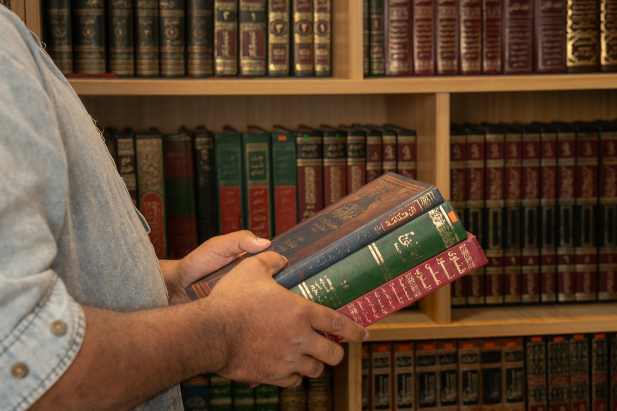 A person holding three hardcover books in front of a wooden library shelf